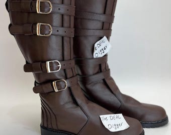 May include: A pair of dark brown mid-calf boots featuring four horizontal straps with silver-toned metal buckles on the outer side. The boots have a rounded toe, a thick black lug sole, and a paper tag attached with the text The DEAL Digger.