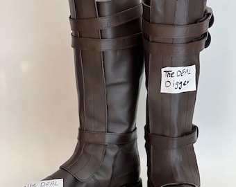 May include: A pair of knee-high dark brown boots featuring a smooth finish, thick black lug soles, and two adjustable buckled straps around the upper shaft and ankle. Each boot displays a white paper label with the text The DEAL Digger.