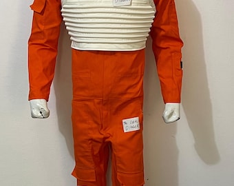 May include: A bright orange jumpsuit with long sleeves and multiple cargo pockets, worn with a white ribbed chest protector. The garment features small white labels with the handwritten text The DEAL DIGGER attached to the chest, thigh, and leg.