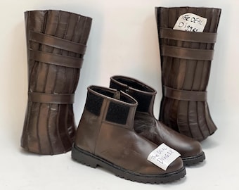 May include: A pair of brown ankle boots with black velcro closures and a pair of matching tall brown leg guards featuring vertical panels and horizontal straps. A white tag with the text The Deal Digger is attached to the boots and one guard.