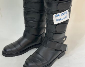 May include: A pair of black mid-calf boots featuring a quilted upper design, multiple horizontal hook-and-loop fastener straps, and a thick lug sole. A white tag with the handwritten text THE DEAL DIGGER is attached to the side of one boot.