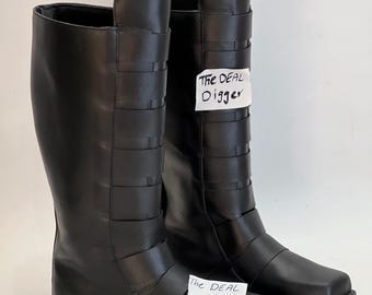 May include: A pair of black mid-calf boots featuring a smooth finish and a series of horizontal straps across the front. A white tag with the handwritten text "The DEAL Digger" is attached to the shaft and the toe area of the boots.