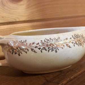 Peut inclure: Une saucière en céramique de couleur crème ornée d'un motif floral doré délicat sur l'extérieur. Le récipient possède une anse unique sur le côté gauche, un large bec verseur sur la droite et une finition émaillée craquelée visible.