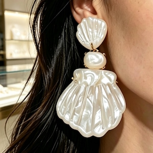 May include: A statement earring featuring a large, white, pearlescent shell-shaped pendant suspended from a smaller, smooth, round, off-white bead, which is attached to a top shell-shaped piece, all connected by small gold-toned metal links.
