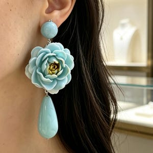 Peut inclure: Une boucle d'oreille pendante composée d'une sphère bleu clair lisse en haut, d'une grande fleur bleu clair à plusieurs couches avec un centre doré au milieu, et d'un pendentif lisse en forme de goutte d'eau bleu clair suspendu en bas.