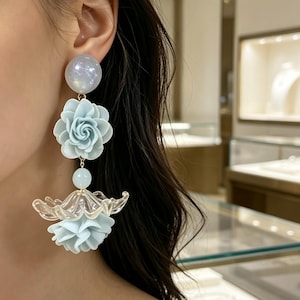 Puede incluir: Un pendiente largo con un poste redondo iridiscente, un componente floral azul claro, una pequeña cuenta redonda, un elemento transparente en forma de pétalo y una pieza floral azul claro en la parte inferior, dispuestos verticalmente.