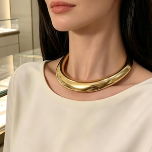 May include: A thick, polished gold-tone choker necklace with a smooth, rounded, tubular design that sits closely around the base of the neck. The metal surface is highly reflective, creating a bright, uniform metallic sheen across the entire piece.