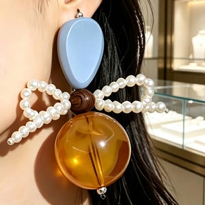 Puede incluir: Un pendiente llamativo que presenta una pieza superior lisa en forma de lágrima de color azul claro, una cuenta central de madera marrón veteada, un lazo hecho de perlas blancas y una cuenta inferior esférica, translúcida y de color ámbar.