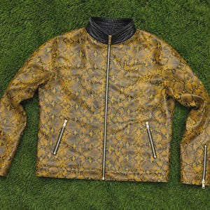Puede incluir: Una chaqueta de manga larga con un patr&oacute;n de piel de serpiente en color amarillo mostaza y negro. La chaqueta tiene un cuello alto negro, una cremallera frontal plateada de cuerpo entero y cuatro bolsillos con cremallera plateada en el pecho y pu&ntilde;os.
