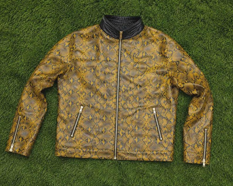 Puede incluir: Una chaqueta de manga larga con un patr&oacute;n de piel de serpiente en color amarillo mostaza y negro. La chaqueta tiene un cuello alto negro, una cremallera frontal plateada de cuerpo entero y cuatro bolsillos con cremallera plateada en el pecho y pu&ntilde;os.