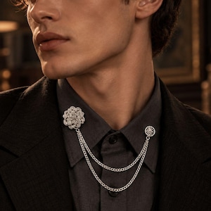 May include: A silver-toned collar brooch featuring a textured, circular floral-shaped pin on one side and a smaller, round, sunburst-patterned pin on the other, connected by two delicate, draped silver-toned metal chains across the shirt collar.