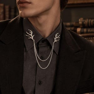 Puede incluir: Un alfiler de cuello plateado con dos adornos simétricos en forma de cornamenta, unidos por una fina cadena metálica de doble capa. El accesorio está sujeto a las puntas de una camisa gris oscuro que se lleva bajo una americana oscura.