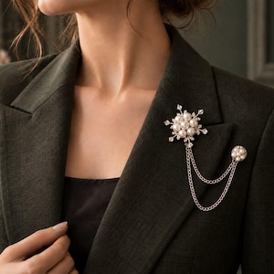 May include: A silver-toned brooch featuring a central snowflake design adorned with white pearls. Two delicate silver chains connect the snowflake to a smaller, circular pearl-encrusted pin, all attached to a dark olive green blazer.