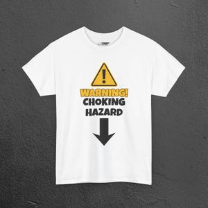 Puede incluir: Una camiseta blanca de manga corta con una señal de advertencia triangular amarilla con un signo de exclamación, seguida del texto WARNING! CHOKING HAZARD en letras negras y amarillas, con una gran flecha negra apuntando hacia abajo.