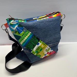 May include: A crossbody bag constructed from blue denim fabric featuring a diagonal panel of multicolored geometric patterned fabric. The bag includes a black adjustable strap, silver metal hardware, and a top zipper closure.
