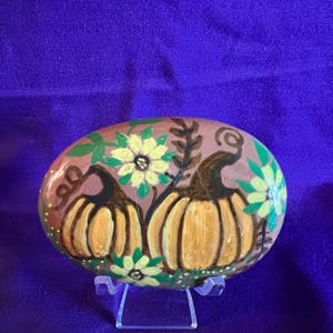 May include: An oval-shaped stone painted with two orange pumpkins featuring dark brown stems and vines, surrounded by light yellow flowers and green leaves on a muted mauve background, displayed on a clear plastic stand.