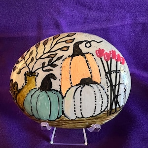 May include: A smooth, oval-shaped stone painted with a seasonal scene featuring three pumpkins in shades of orange, light blue, and white. The design includes a yellow vase with brown branches, a cluster of pink flowers, and black outlines.