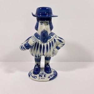 May include: A ceramic figurine of a person wearing a wide-brimmed hat, a ruff collar, and a striped doublet. The figure is decorated in blue and white with a beard and stands on a circular base featuring the text P E N E J 682 in blue ink.