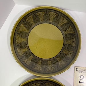 May include: A round ceramic plate featuring a solid olive green centre surrounded by a wide border decorated with a dark brown repeating pattern of stylised leaf shapes and geometric motifs. A portion of a game card is visible in the corner.