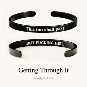 May include: Two black metal cuff bracelets with open ends. The top bracelet features the white engraved text "This too shall pass" and the bottom bracelet features the white engraved text "BUT FUCKING HELL". Below the bracelets is the text "Getting Through It" and "Barely, but still".