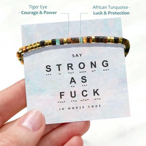 May include: A beaded bracelet featuring alternating brown tiger eye, turquoise, and gold-toned cylindrical beads. The bracelet is displayed on a card with the text "SAY STRONG AS FUCK IN MORSE CODE" and labels identifying the stone types.