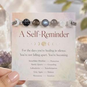 May include: A beaded bracelet featuring a snowflake obsidian, smoky quartz, labradorite, gray agate, and moonstone bead, flanked by two silver crescent moon charms. The bracelet is displayed on a card titled A Self-Reminder with text.