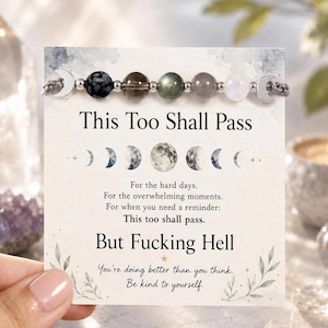 May include: A beaded bracelet featuring a row of round stones in black, brown, green, grey, and white, flanked by silver crescent moon charms. The bracelet is attached to a card printed with moon phases and the text This Too Shall Pass and But Fucking Hell.