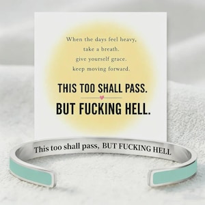 May include: A silver-toned metal cuff bracelet with a light blue enamel inlay. The bracelet is engraved with the text: This too shall pass, BUT FUCKING HELL. It is displayed in front of a white card featuring the same text and a yellow circle.