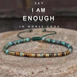 May include: A beaded bracelet featuring a mix of turquoise-colored stone beads, brown tiger eye beads, and metallic gold-tone cylindrical spacers. The bracelet has a dark green adjustable cord closure and displays the text SAY I AM ENOUGH IN MORSE CODE.