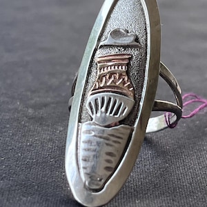 May include: An elongated oval silver ring featuring a textured gray background. The center of the ring displays a metallic inlay depicting a stylized vessel or pot with horizontal and vertical line patterns, topped by a small cloud-like shape.