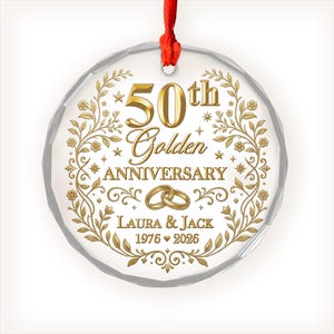 May include: A circular clear glass ornament with a red ribbon hanger, featuring gold text that reads 50th Golden ANNIVERSARY, LAURA & JACK, and 1976 2026. The design includes gold floral vines, stars, and two interlocking gold wedding rings.