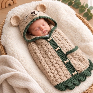 Puede incluir: Una manta de arrullo para bebé tejida en crochet beige con capucha, orejas de oso y ribete verde oscuro. La manta tiene una abertura frontal con cuatro botones en forma de hueso y un pequeño bolsillo lateral con borde festoneado.