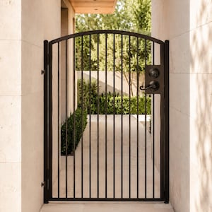 May include: A black metal gate with a curved top and vertical bars, installed between two light-colored stone walls. The gate features a black handle and a circular lock mechanism on the right side, providing a clear view through the vertical bars.