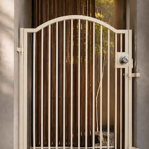 May include: A cream-colored metal gate featuring a vertical bar design with an arched top rail. The gate is equipped with a circular silver-toned knob and a latch mechanism, mounted between two concrete walls with visible hinges on the right side.
