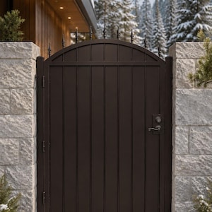 May include: A dark brown metal gate with a vertical panel design and an arched top. The gate features a series of pointed finials along the upper edge and a black handle with a locking mechanism on the right side, mounted between two stone pillars.