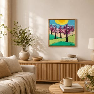 Op de afbeelding: Een vierkant schilderij op canvas in een lichthouten lijst met een landschap van roze bloeiende bomen, een heldere gele zon en glooiende groene heuvels onder een blauwe lucht. Het kunstwerk toont donkere boomstammen en roze bloesems.