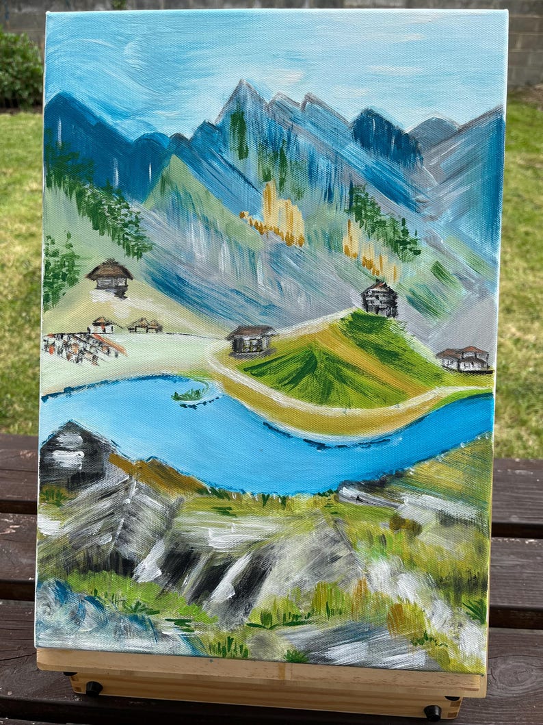 May include: An acrylic painting on canvas depicting a mountainous landscape with a winding blue river, green grassy hills, and several small dark-roofed cabins. The background features jagged blue and grey mountain peaks under a light blue sky.