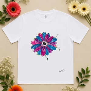 May include: A white short-sleeve t-shirt featuring a central graphic of a stylized flower with vibrant blue and magenta petals and a dark center. The design includes a small signature that reads "Mimla P" in the lower right area of the shirt.