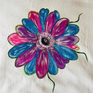 May include: A hand-painted illustration of a flower on a white fabric surface. The flower features alternating petals in vibrant shades of blue, purple, and pink, with a textured, dark center and thin, green, hand-drawn stems extending outward.