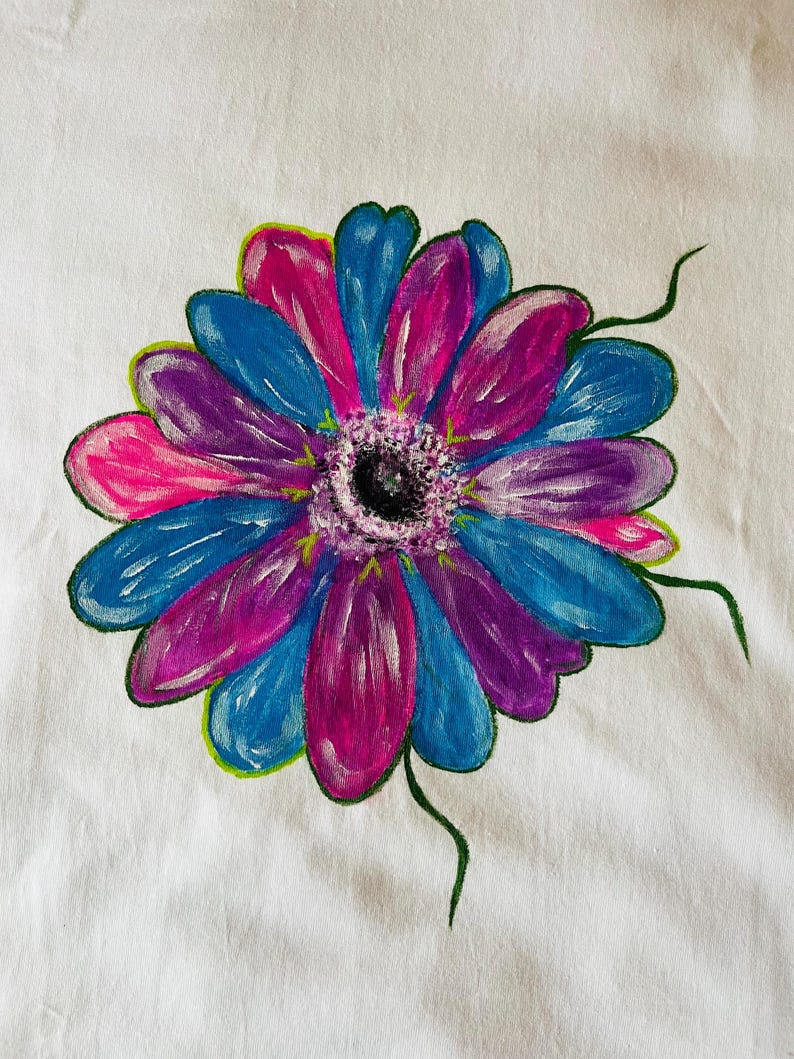 May include: A hand-painted illustration of a flower on a white fabric surface. The flower features alternating petals in vibrant shades of blue, purple, and pink, with a textured, dark center and thin, green, hand-drawn stems extending outward.
