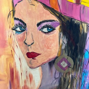 Peut inclure: Une peinture acrylique sur toile repr&eacute;sentant le portrait d'une personne aux yeux bleus, aux cheveux fonc&eacute;s et aux l&egrave;vres rouges. Le sujet porte une coiffe rose et jaune ainsi qu'une boucle d'oreille circulaire avec un losange violet et une croix blanche.