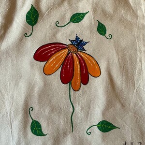 May include: A beige fabric tote bag featuring a hand-painted design of a flower with alternating red and orange petals, a dark blue butterfly with white dots resting on the center, and four green leaves. The signature Mirela P. is in the corner.