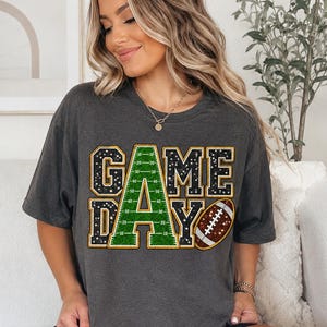 May include: A charcoal gray short-sleeve t-shirt featuring the text "GAME DAY" in a bold, block-letter style. The letters are filled with a green football field pattern and black glitter, while a brown football graphic sits to the right.