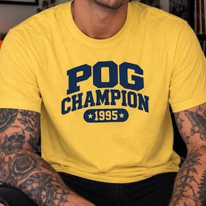 May include: A bright yellow short-sleeved t-shirt featuring the dark blue text POG CHAMPION centered on the chest, with the year 1995 enclosed between two stars below the text in a matching dark blue font and a classic crew neckline.
