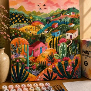 Op de afbeelding: Een schilderen-op-nummer set met een canvas waarop een woestijnlandschap met cactussen, heuvels en kleine huisjes onder een roze lucht staat afgebeeld. Inclusief vierentwintig genummerde verfpotjes en drie penselen voor het schilderen.