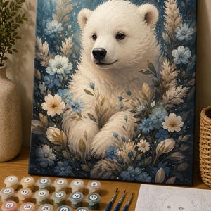 Op de afbeelding: Een schilderen-op-nummer set met een canvas van een witte ijsberenwelp omringd door blauwe en witte bloemen. De set bevat een strip met genummerde verfpotjes en drie penselen, samen met een gedrukt instructieblad voor de afbeelding.