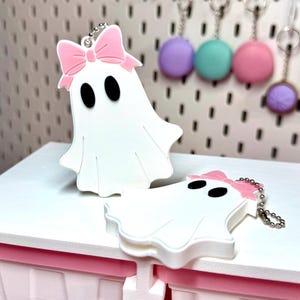 May include: Two white ghost-shaped keychains featuring black oval eyes and a light pink bow attached to the top. Each ghost figure includes a silver-toned metal ball chain for attachment, with one ghost standing upright and the other lying flat.