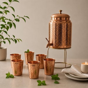 May include: A hammered copper water dispenser with a lid and a spigot, resting on a gold-toned metal stand, accompanied by five matching hammered copper tumblers. The set is arranged with small green leaves scattered around the base.