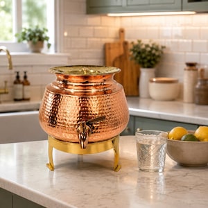 May include: A hammered copper beverage dispenser with a polished gold-tone lid and matching gold-tone metal stand. The dispenser features a front-mounted spigot for liquid dispensing, showcasing a rounded, wide-body design with a metallic finish.