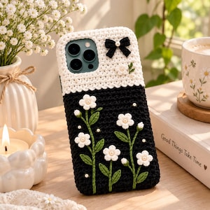 Può includere: Una custodia per telefono rettangolare con un design all'uncinetto bicolore, caratterizzata da una sezione superiore bianca e una inferiore nera. La custodia è decorata con un piccolo fiocco nero, fiori bianchi all'uncinetto e steli verdi.